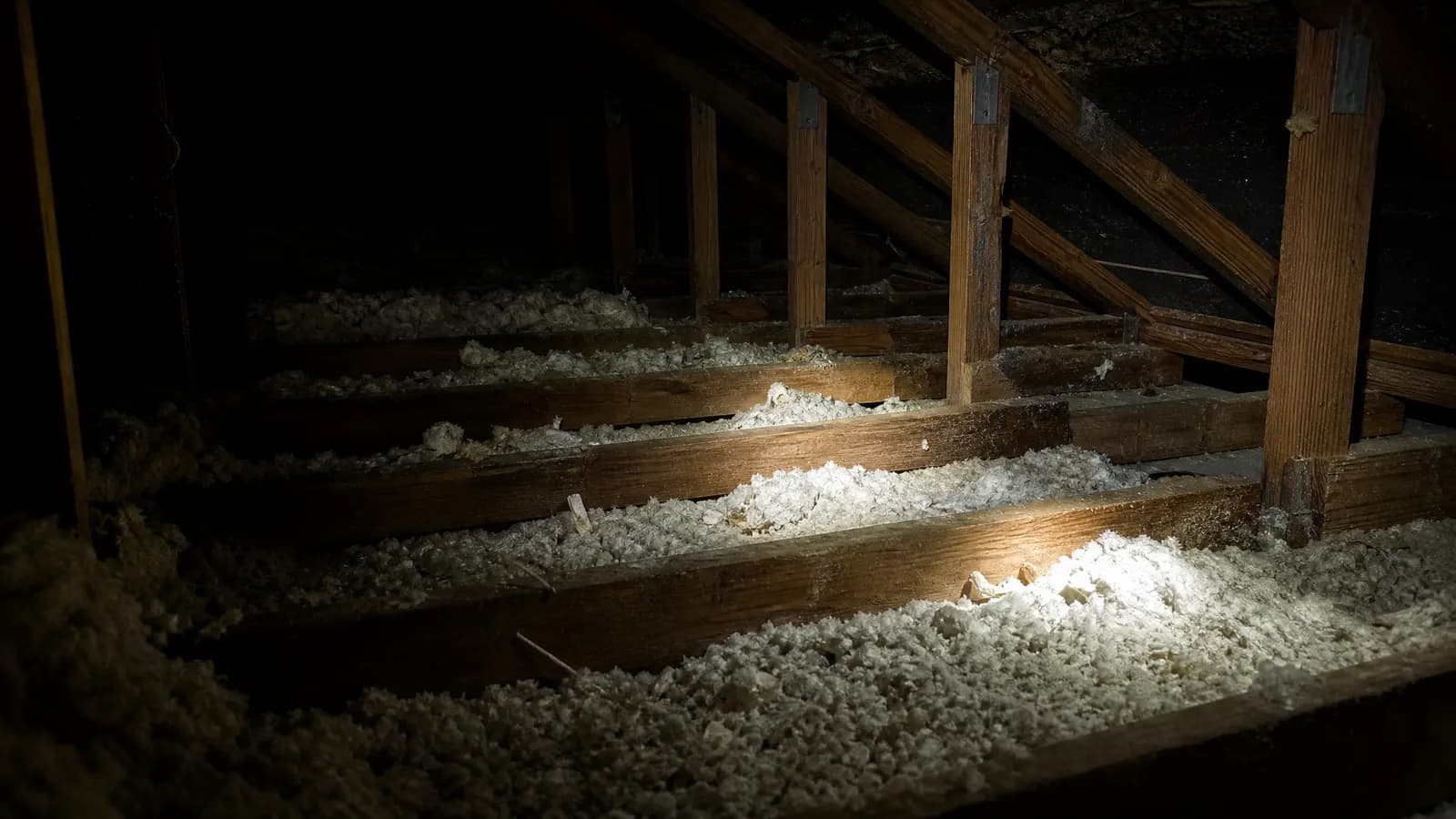 Dark residential attic at night with rafters and insulation lit by a single flashlight beam