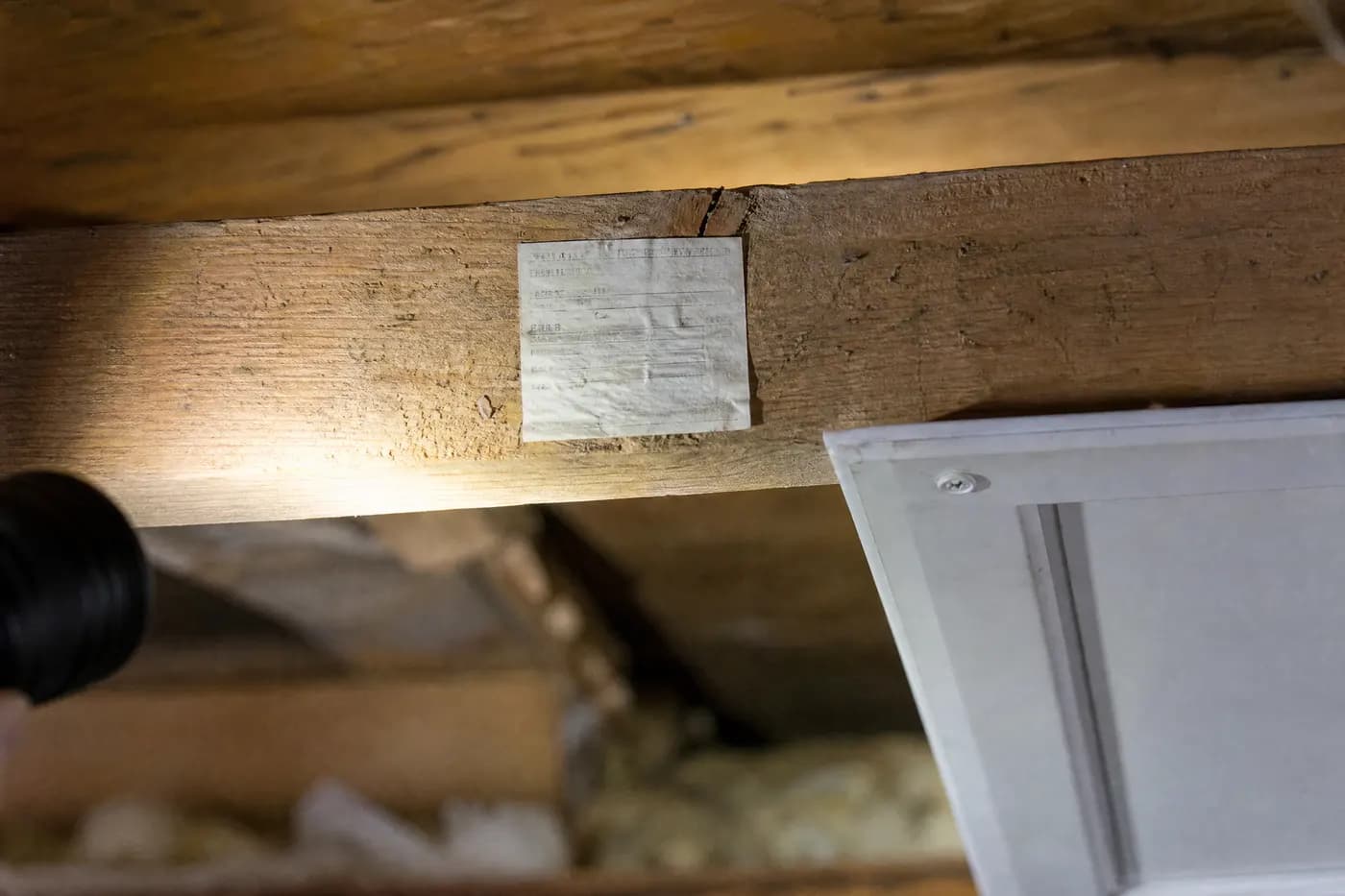 Weathered inspection notice sticker fixed to a wooden attic joist beside the hatch, its printed text no longer legible