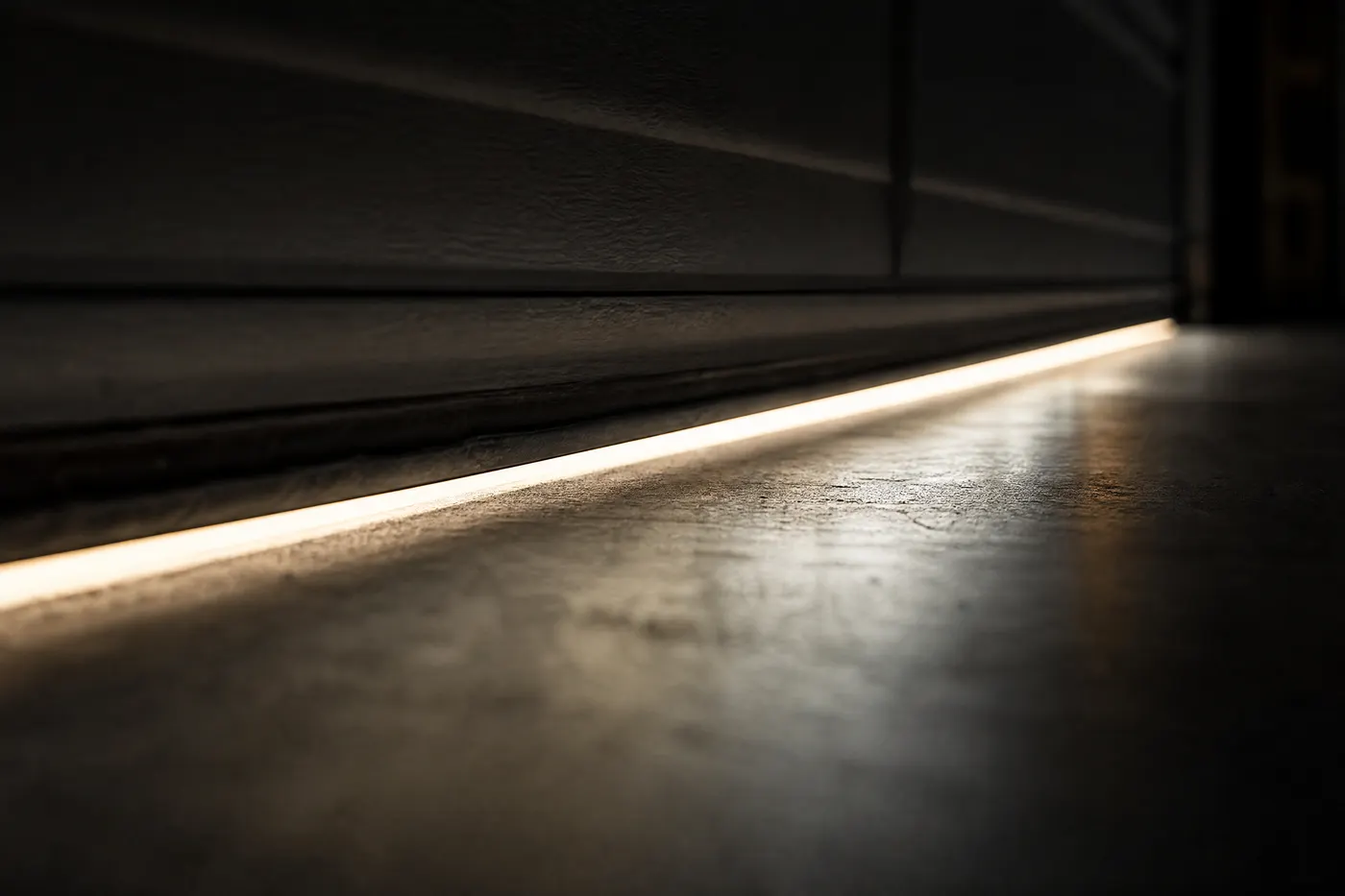 View from inside a dark garage at night of a thin line of outside light glowing through the gap beneath the flattened rubber bottom seal of a closed garage door