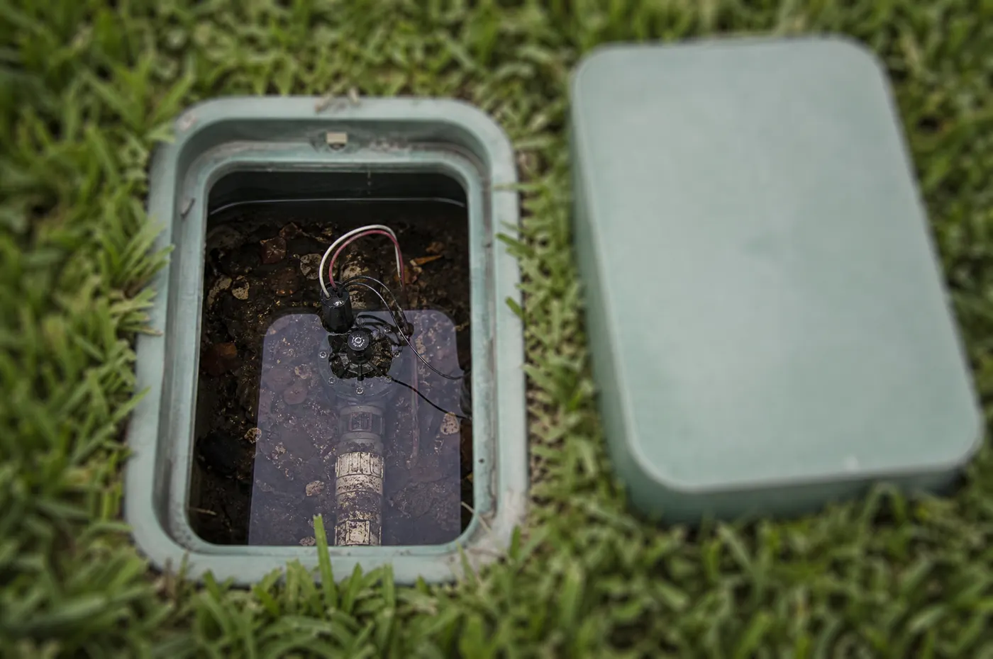 Green plastic irrigation valve box in a lawn with its lid lifted off, showing dark standing water in the bottom of the box around the valve